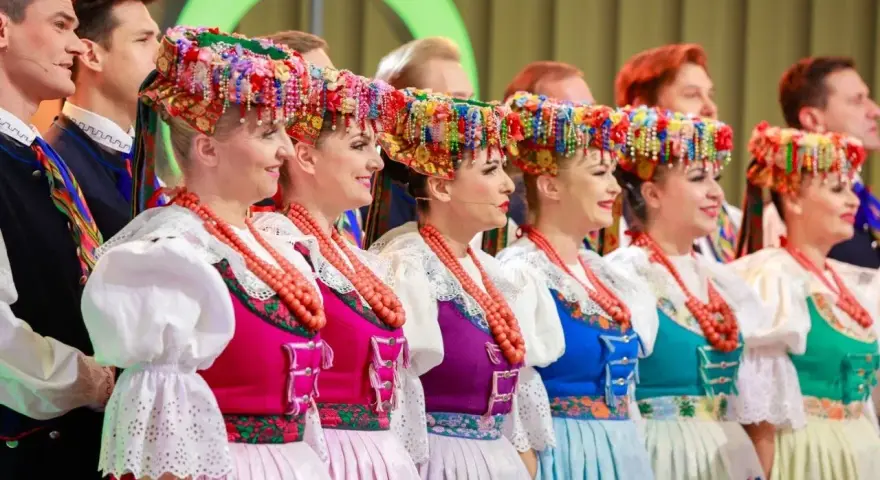 Szła dzieweczka w tradycyjnym stroju, z barwnym wiankiem na głowie, obok innych w ludowych strojach.