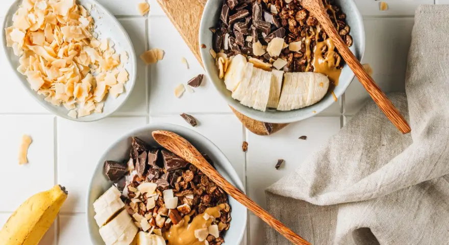 Due ciotole con granola fatta in casa, banana a fette, cioccolato fondente e burro d'arachidi. Cocco in scaglie a lato.