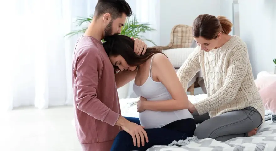 Mężczyzna wspiera ciężarną kobietę, a doula delikatnie masuje jej plecy.