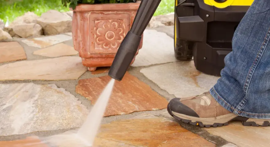 Pulire pavimento esterno con idropulitrice. Un getto d'acqua potente rimuove lo sporco dalle pietre del patio, mentre una persona in jeans e scarponi osserva.