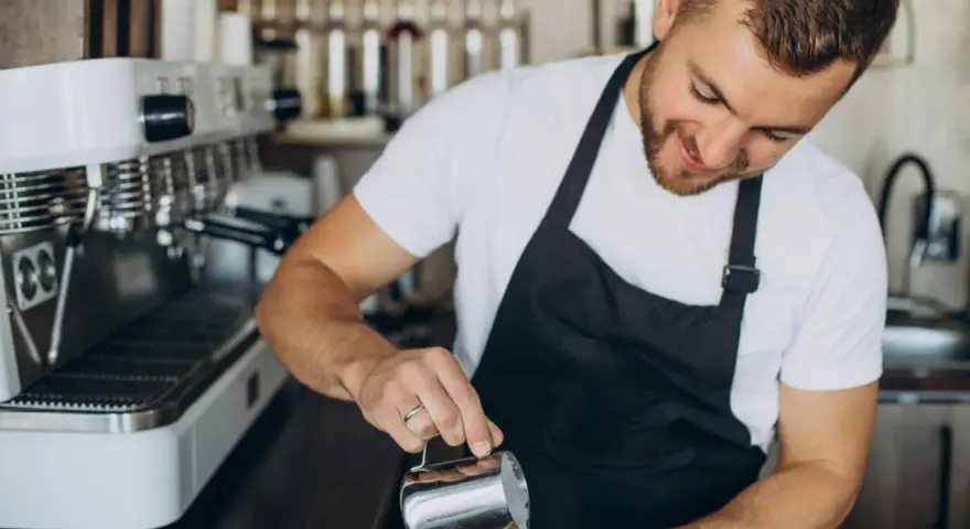 Uśmiechnięty barista w czarnym fartuchu nalewa mleko do kawy. Obok stoi profesjonalna ekspres do kawy.
