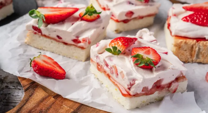 Saftige Stücke eines Erdbeerkuchen Blechs mit frischen Erdbeeren und Sahne, auf Backpapier angerichtet.