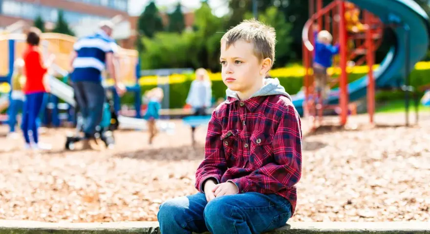 Chłopiec z zespołem Aspergera siedzi samotnie na placu zabaw, podczas gdy inne dzieci się bawią.