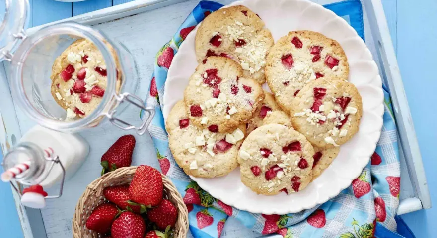 Saftige Erdbeer-Cookies mit weißen Schokostückchen auf einem Teller, daneben frische Erdbeeren und ein Glas Milch.