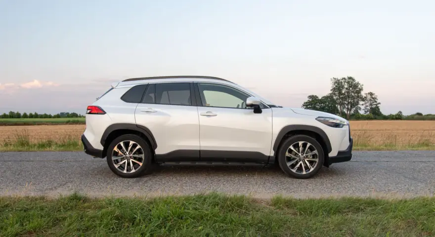 Biała Toyota Corolla Cross Hybrid stoi na poboczu drogi, otoczona polami i zieloną trawą.