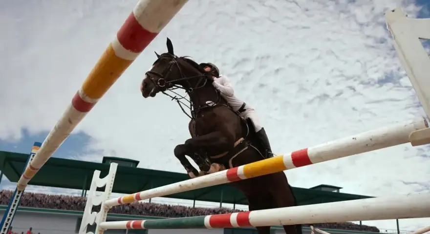 Un cheval brun et son cavalier en blanc effectuent un spectaculaire saut d'obstacles, le tout sous un ciel nuageux.