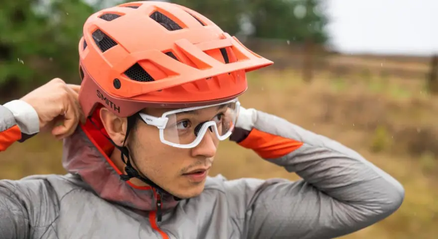 Uomo con casco da bici arancione e occhiali bianchi, mentre regola il casco. Utile per capire come misurare la testa per il casco.
