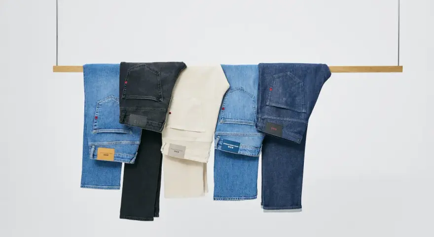 Cinco pares de pantalones vaqueros de distintos tonos cuelgan de una barra de madera. El **denim material** se presenta en azul claro, negro, beige, azul medio y azul oscuro.