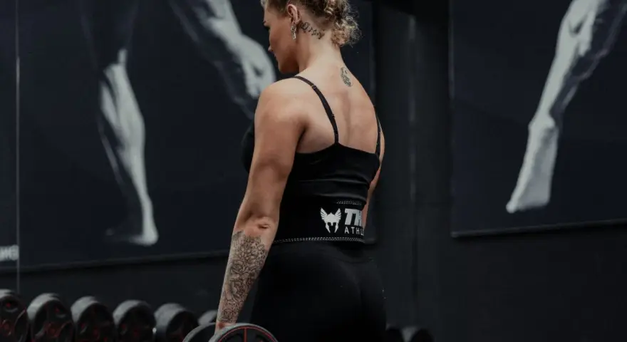 Kvinna med tatueringar utför shrugs med hantlar i ett gym.