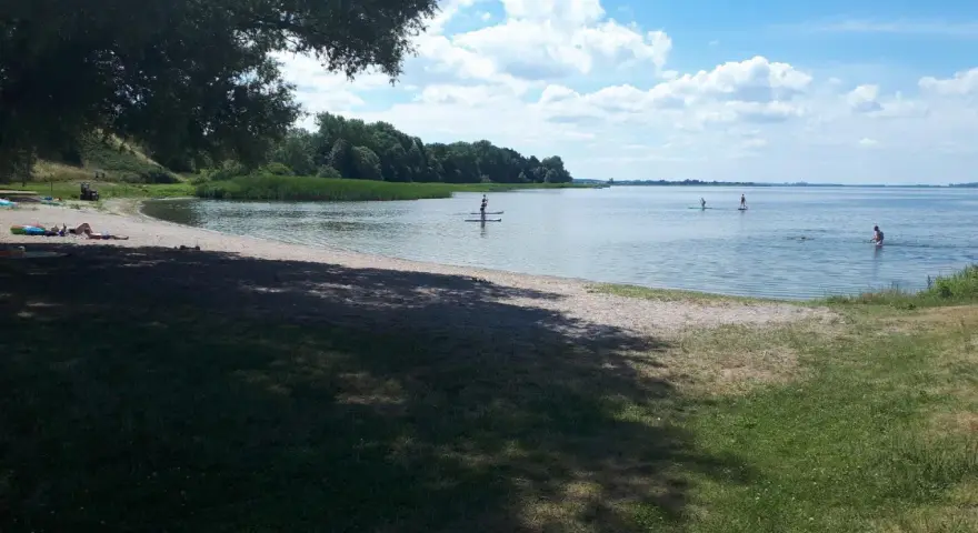 Sonniger Tag am Kummerower See. Menschen entspannen am Strand, andere paddeln auf SUPs. Herrlicher See- und Waldblick.