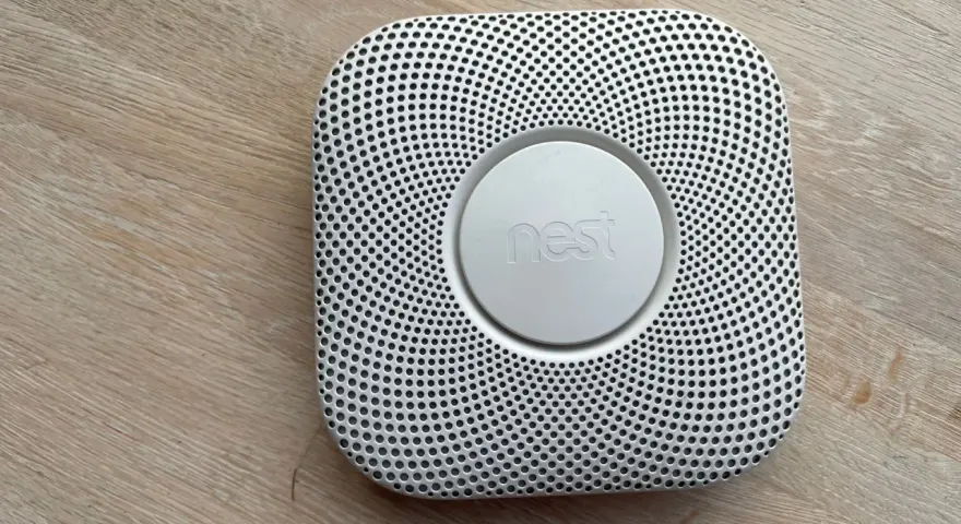 En vit Google Nest Protect-rökdetektor med ett runt "nest"-märke i mitten, placerad på ett trägolv.