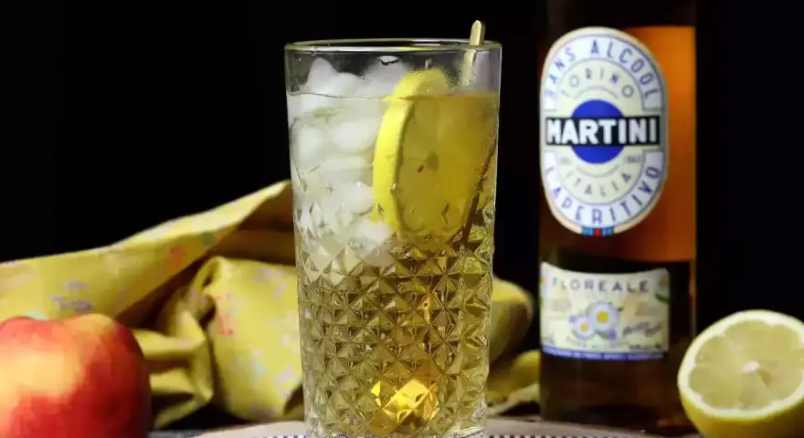 Orzeźwiający drink z Martini i lodem, ozdobiony plasterkiem cytryny. Obok jabłko i butelka Martini.
