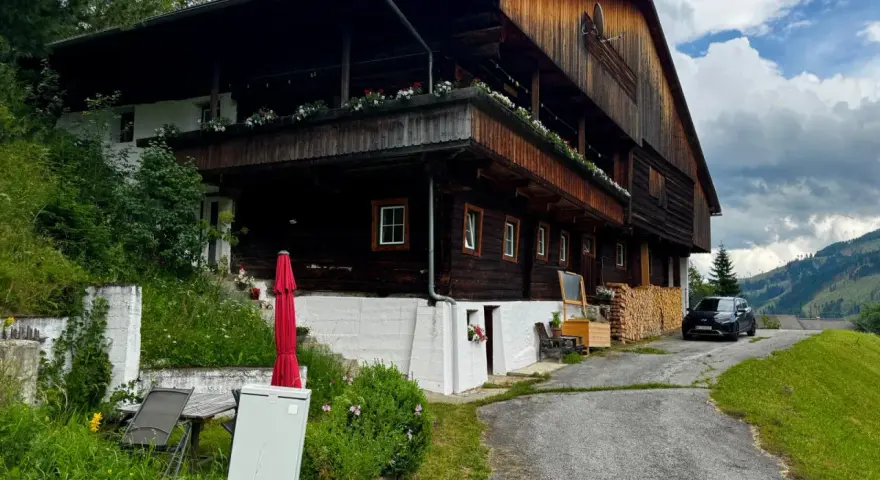 Drewniana austeria w górach, z balkonami ozdobionymi kwiatami. Obok stoi czarny samochód, a w oddali widać zalesione wzgórza.