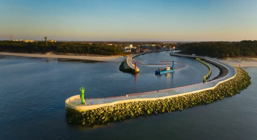 Port w Mrzeżynie z falochronami i latarniami morskimi. Widoczna łódź pogłębiarka. Mrzeżyno atrakcje dla miłośników morza.
