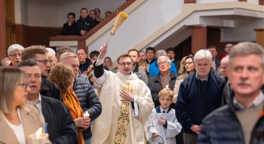 Kapłan błogosławi wiernych podczas liturgii Wielkiej Soboty, unosząc kropidło. W tłumie widać ludzi z zapalonymi świecami.