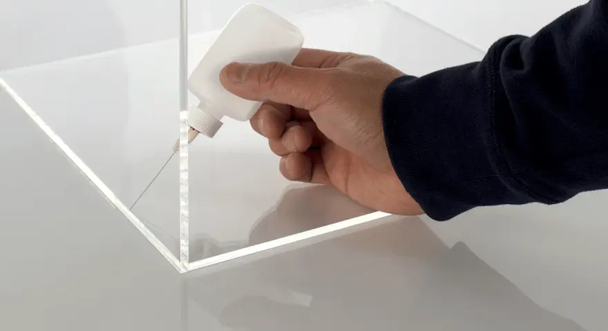 A hand applies adhesive to the corner of a clear plastic box, demonstrating precise gluing of plastic for a clean finish.