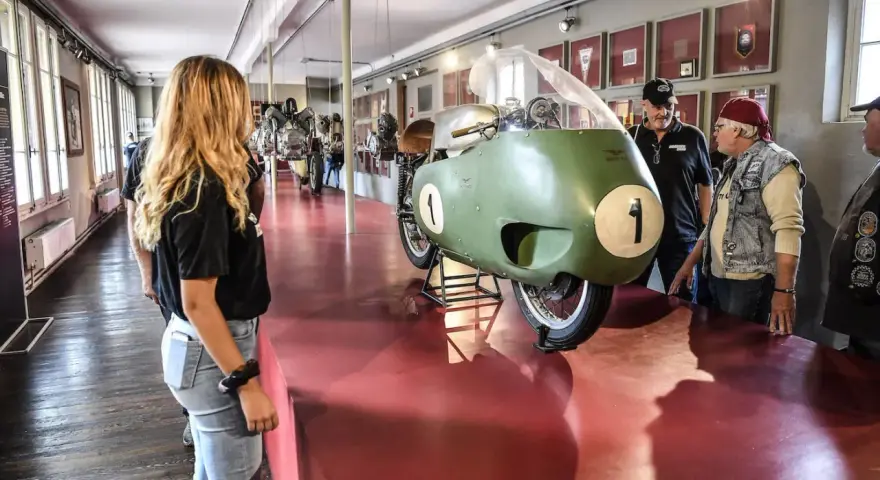 Un raduno moto Guzzi in un museo. Una moto da corsa verde con il numero 1 in primo piano, ammirata da visitatori.