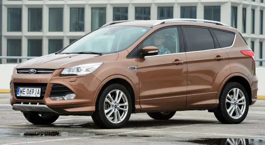 Brązowy Ford Kuga 2019 z kierowcą w okularach, zaparkowany na mokrym parkingu przed nowoczesnym budynkiem.