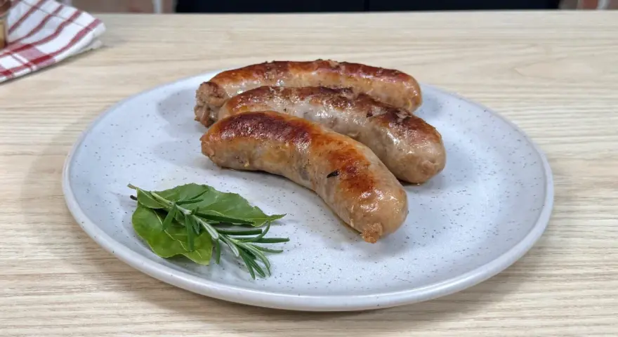 Tre salsicce dorate e succulente, appena cotte in padella, riposano su un piatto con rametti di rosmarino e alloro.