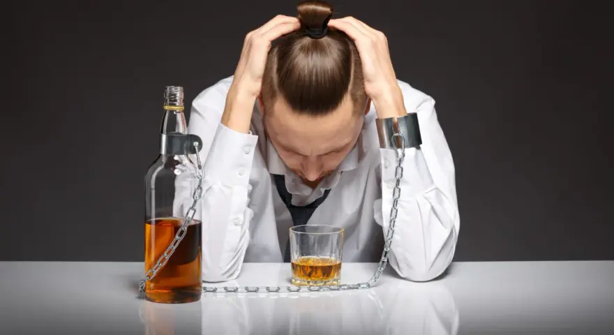 Mężczyzna w kajdanach, przykuty łańcuchem do butelki i szklanki z alkoholem. To symbol zniewolenia przez grupę alkoholową.