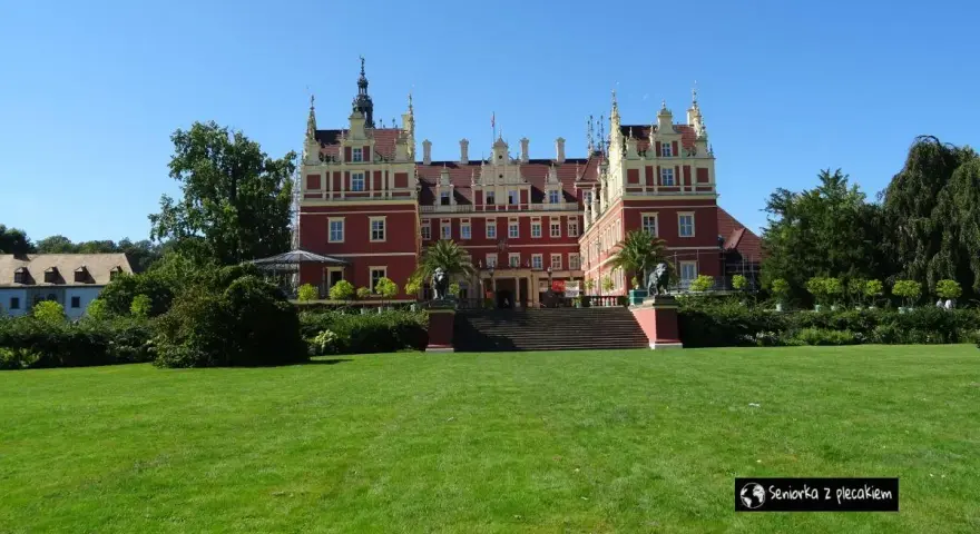 Majestatyczny pałac w parku muzakowskim, otoczony zielonym trawnikiem i drzewami, pod błękitnym niebem.