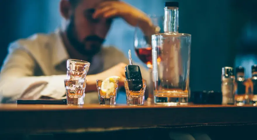 Ein Mann mit Kopfschmerzen sitzt am Tresen, neben Autoschlüsseln in einem Schnapsglas. Alkohol am Steuer unter 21 ist eine Gefahr.