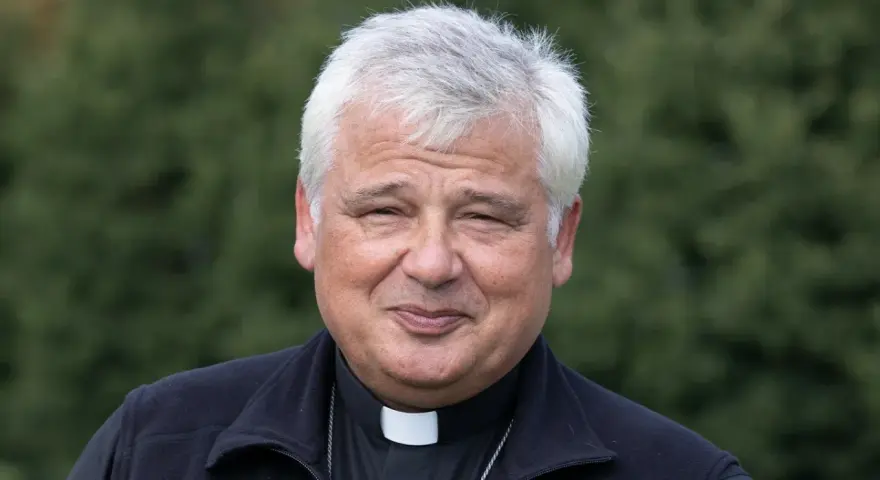 Uśmiechnięty jałmużnik papieski w sutannie i czarnym polarze, na tle zieleni.
