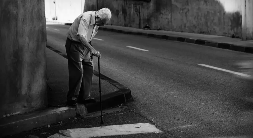 Une photo dans la rue : un vieil homme se penche sur sa canne, le regard perdu, au bord d'une route déserte.