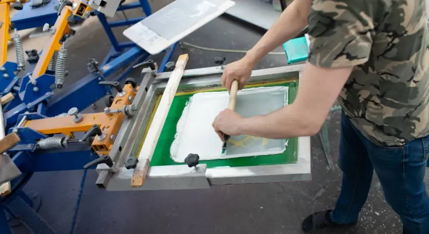 Proceso de serigrafía que es: un operario aplica tinta blanca sobre una pantalla para estampar.