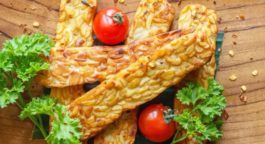 Złocisty, smażony tempeh, czyli co to jest? Podany z pomidorkami i natką pietruszki na drewnianej desce.
