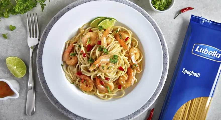 Pyszny makaron z krewetkami na winie, udekorowany limonką i natką pietruszki. Obok opakowanie makaronu spaghetti Lubella.