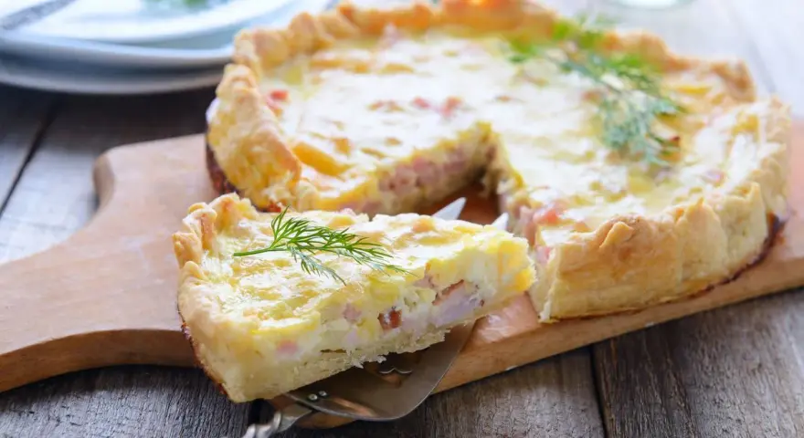 Una fetta di torta salata prosciutto e formaggio, guarnita con aneto, su un tagliere di legno.
