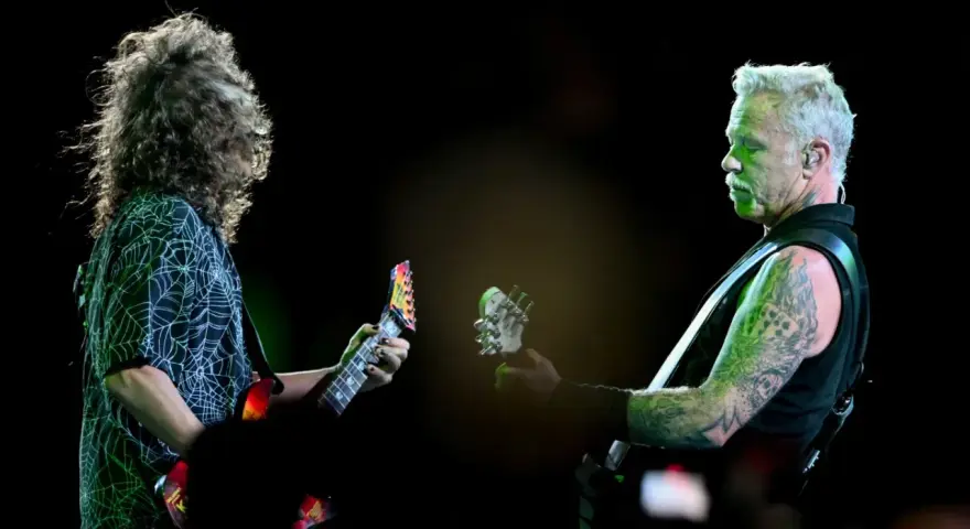James Hetfield, z charakterystycznym wąsem i wytatuowanymi ramionami, gra na gitarze. Obok niego drugi muzyk z burzą włosów.