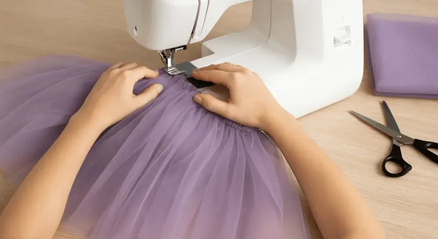 Ręce szyjące fioletową tiulową spódnicę na maszynie. Obok leżą nożyczki i materiał.
