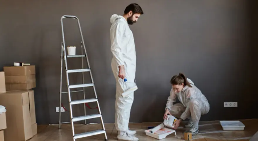 Pareja aprendiendo a pintar su hogar. Él sostiene un rodillo, ella vierte pintura. Hay cajas y una escalera.