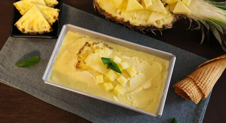 Erfrischendes Ananas Eis in einer Metallform, garniert mit Ananasstücken und Minze. Daneben liegen Ananasspalten und eine Waffel.