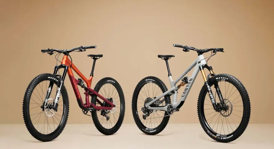 Two full-suspension mountain bikes, one orange and maroon, the other grey, are displayed side-by-side, showcasing the 27.5 vs 29 wheel size debate.