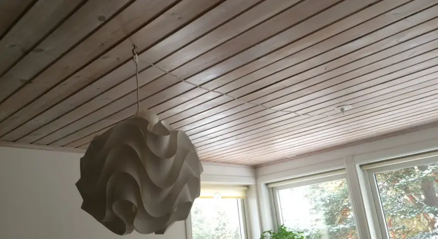 En vit lampskärm hänger från ett tak av träpanel. En tunn sladd dras i taket för att ansluta lampan.