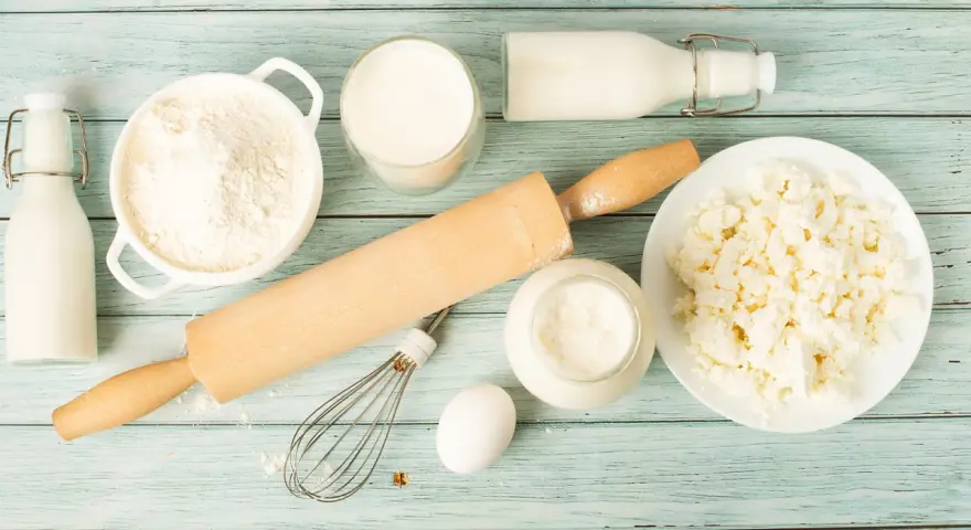 Zutaten für butter ersatz backen: Mehl, Milch, Quark, Ei, Schneebesen und Nudelholz auf blauem Holzuntergrund.