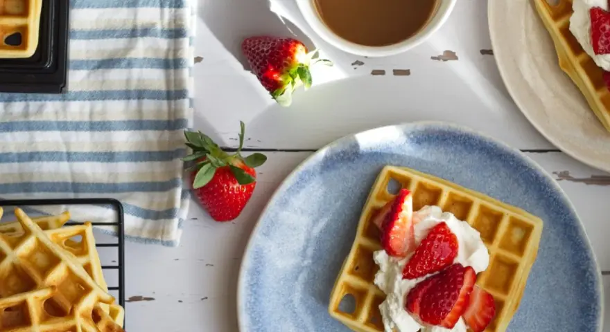 Frische belgische Waffeln mit Erdbeeren und Sahne auf einem blauen Teller, daneben eine Tasse Kaffee und weitere Waffeln auf einem Gitter.