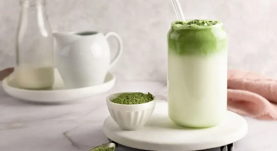 Pyszny matcha latte w szklance, obok miseczka z zielonym proszkiem i łyżeczka.