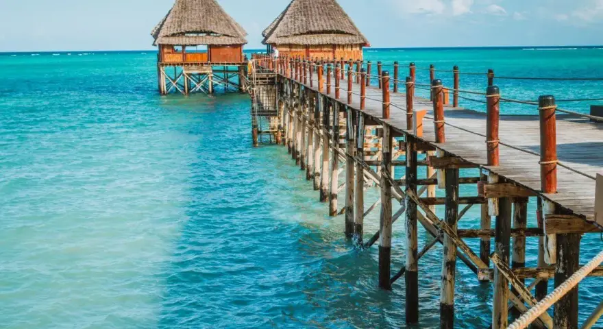 Drewniany pomost prowadzi do chat na palach nad turkusową wodą. Zanzibar, pora deszczowa, ale słońce świeci.