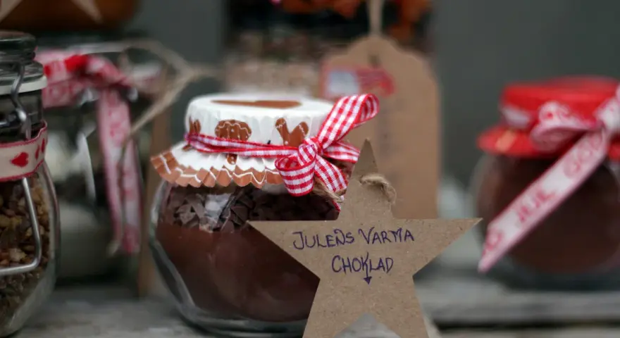 Burkar med ingredienser för varm choklad jul, dekorerade med röda band och pepparkaksfigurer. En stjärna med texten "Julens varma choklad" hänger framför.