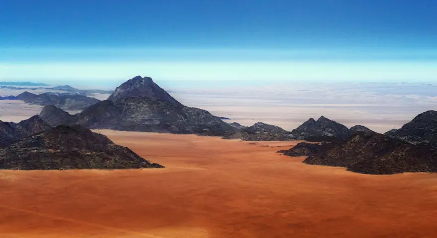 Pustynny krajobraz w **Afryce, Namibia**. Czerwona ziemia i skaliste góry pod błękitnym niebem.