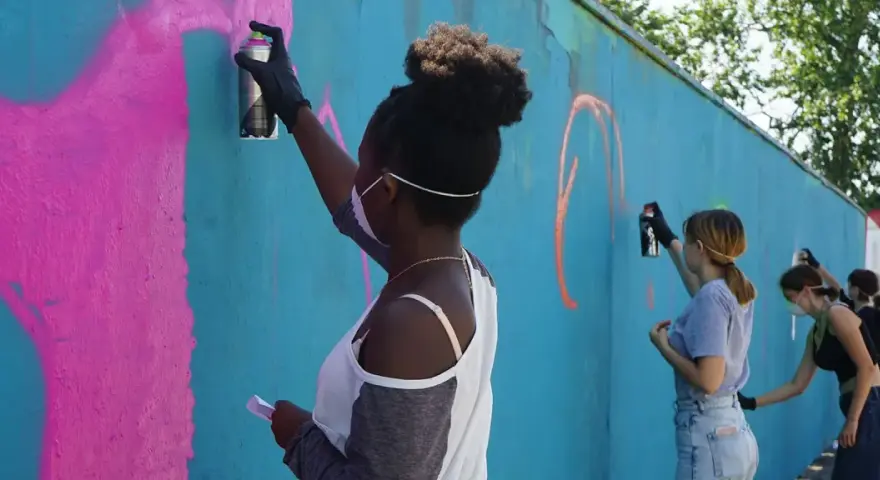 Ungdomar målar graffiti på en blå vägg. En flicka med afrohår sprayar rosa färg.