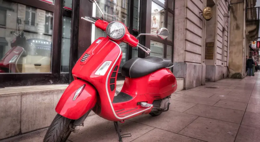 Czerwony skuter Vespa zaparkowany na chodniku przed budynkiem.