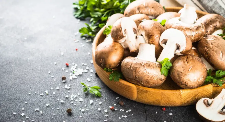 Świeże pieczarki w drewnianej misce z natką pietruszki i solą na ciemnym tle.