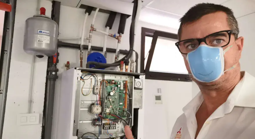 Técnico con mascarilla realiza mantenimiento aerotermia. Equipo abierto, circuito visible. Pulgar arriba.