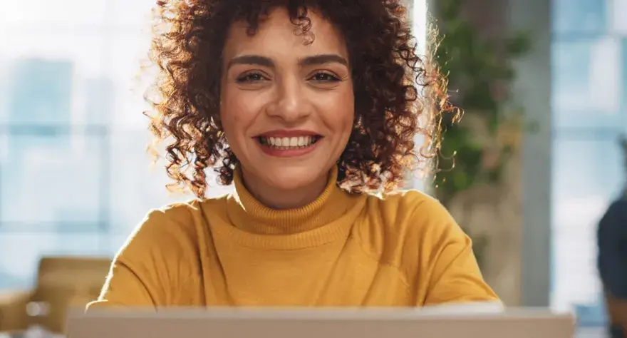 Lächelnde Frau mit Locken am Laptop, bereit für ihre Bewerbung als studentische Hilfskraft.