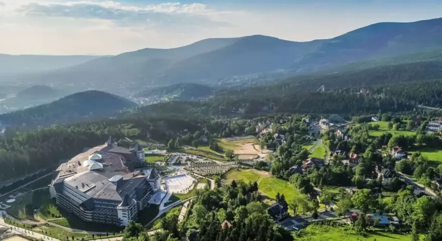 Malownicze Karkonosze latem: rozległy kompleks hotelowy otoczony zielenią, z widokiem na góry i miasteczko. Idealne miejsce na wypoczynek i odkrywanie atrakcji.